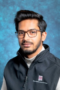 Sarhan Osman Bhuiya- The image shows a person with short dark hair and a well-groomed beard, wearing rectangular eyeglasses, facing the camera. The individual is dressed in a black zip-up vest with a white logo and text over a light-colored turtleneck shirt. The background is a mottled blue texture, providing contrast to the subject's attire. The lighting is even, highlighting the person’s facial features clearly.